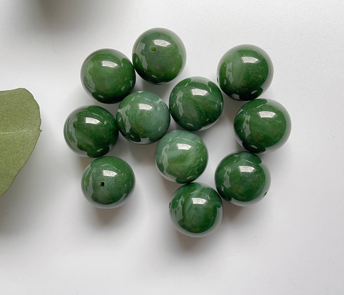Top view of polished green jade beads with drilled holes scattered on white background beside a leaf