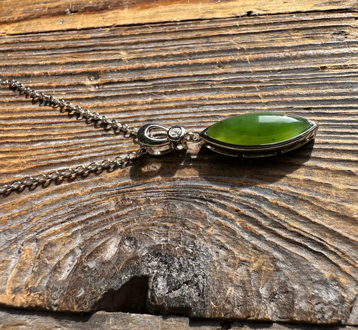 Elongated marquise green jade pendant with a decorative silver setting and chain placed on a rustic wood surface