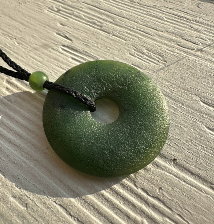 Matte green jade donut pendant with central hole on a black braided cord resting on a light wood surface.