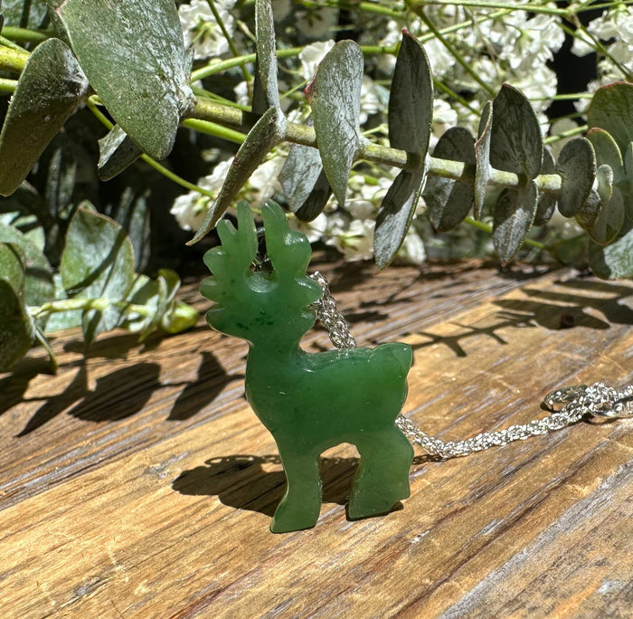 Green nephrite jade deer pendant on a silver chain displayed on a wooden surface with eucalyptus leaves
