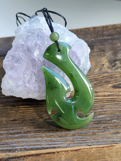 Green nephrite jade fish hook pendant on black cord placed beside a purple amethyst cluster