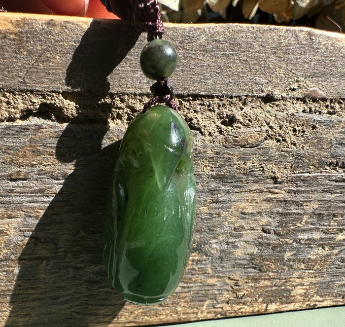 Close up of green jade bird pendant with round bead above on dark braided cord against wood
