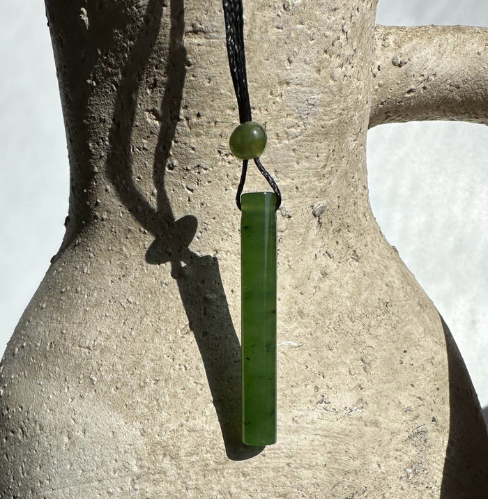 Green jade cylinder pendant on black cord with a round bead above, set against a textured stone backdrop.