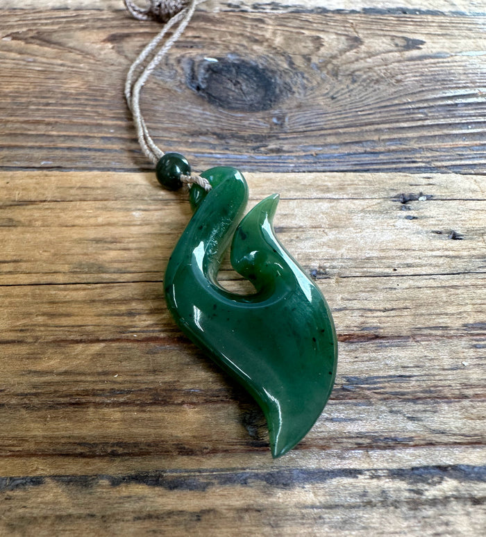 Green jade fish hook pendant with cord and bead resting on a rustic wooden surface