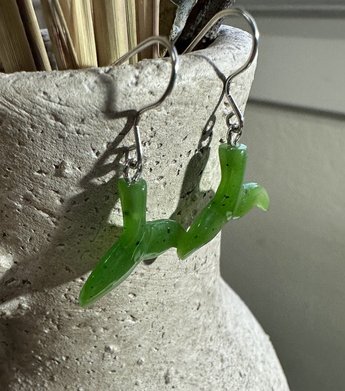 Green jade whale tail earrings with silver hooks hanging from a textured light display