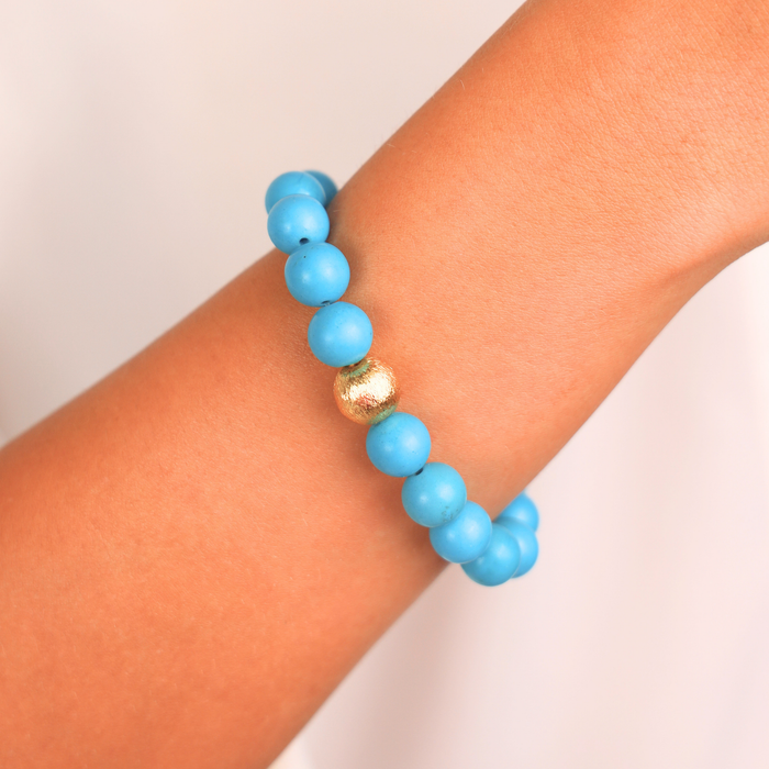 A light blue beaded bracelet with a single textured gold accent bead, shown worn on a person's wrist against a soft off-white background.
