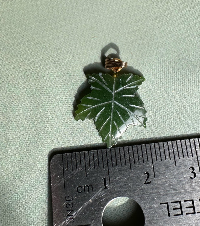 Single dark green maple leaf pendant with gold bail placed on light green fabric beside a ruler