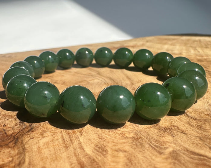 A beaded stretch bracelet made of polished dark green jade round beads, displayed on a textured wooden surface with natural sunlight and shadows.