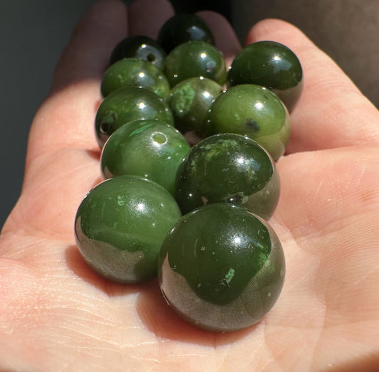 Group of 14mm Canadian Cassiar jade beads held in a hand, polished dark green spheres with drilled holes