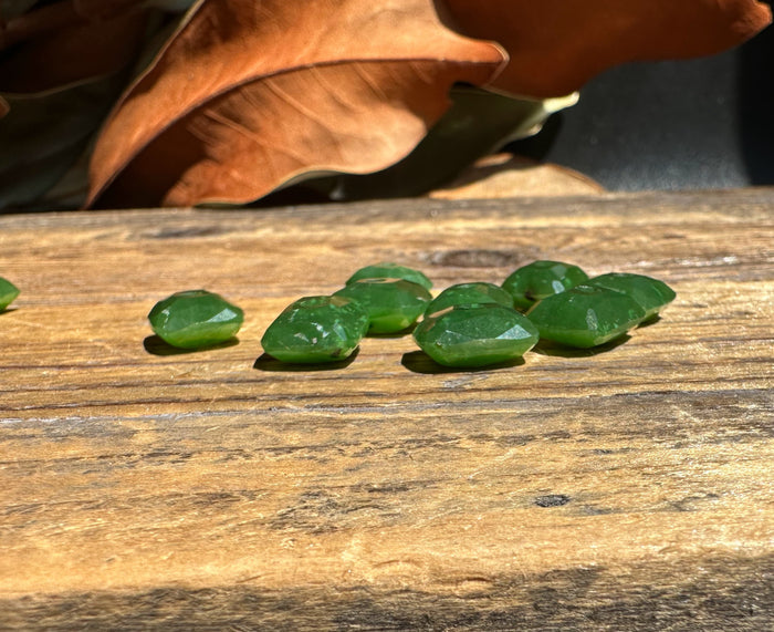 Small green jade beads with faceted surfaces scattered on a weathered wooden surface with a dried leaf in the background