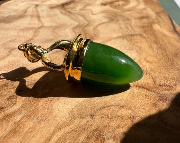 Gold-tone pendant with a green jade drop and ornate bail on a wood surface.