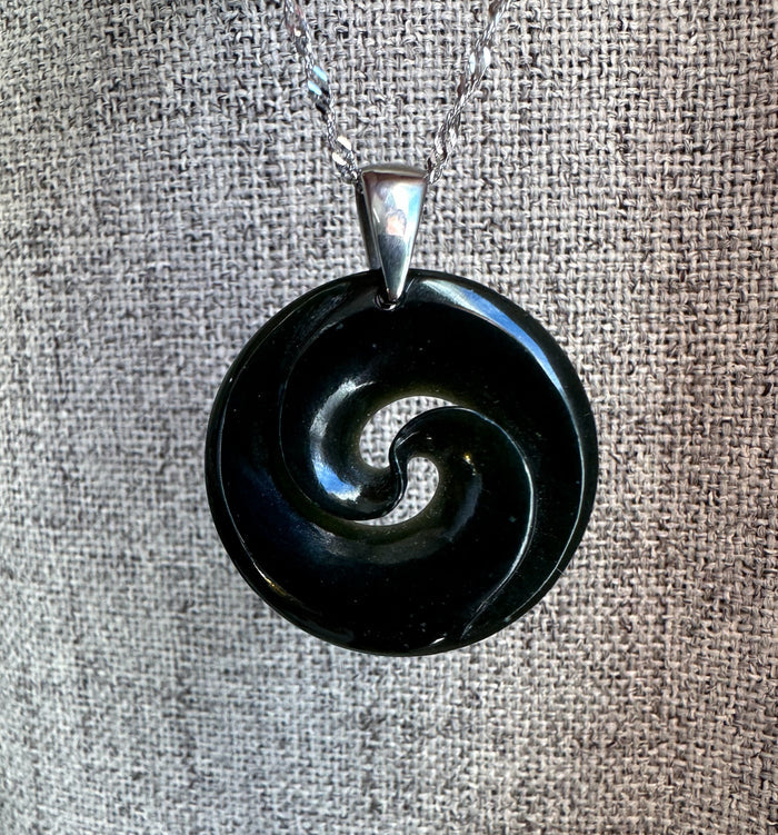 Dark glossy black nephrite jade koru pendant with spiral design and silver bail on a textured gray fabric background.