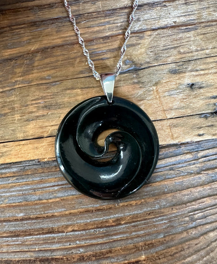 Glossy black nephrite jade koru pendant with swirling spiral resting on a wooden surface.