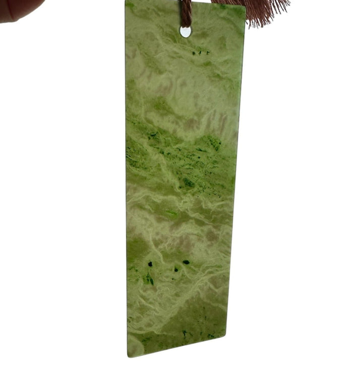 Close-up of green jade bookmark six inches long with brown tassel held by a finger top hole visible against a white background