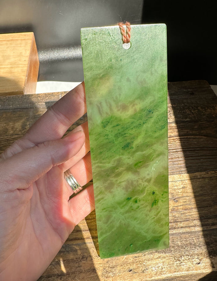 Six inch green jade bookmark being held by a hand, showing a marbled surface and a brown cord at the top.