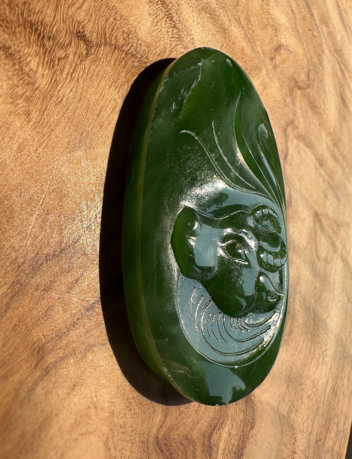 A polished dark green jade pendant featuring a carved buffalo face, displayed on a warm wood grain background.