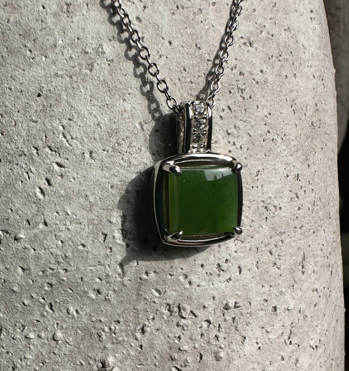 Close-up of green jade pendant in a silver bezel with a studded bail on a textured stone surface.