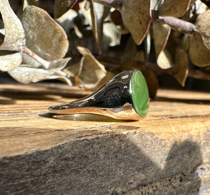 Angled view of a silver ring with a green jade oval on a wooden surface with dried leaves in the background.