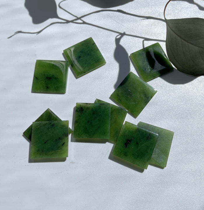 Green jade square cabochons on a white surface with shadows and a dried leaf