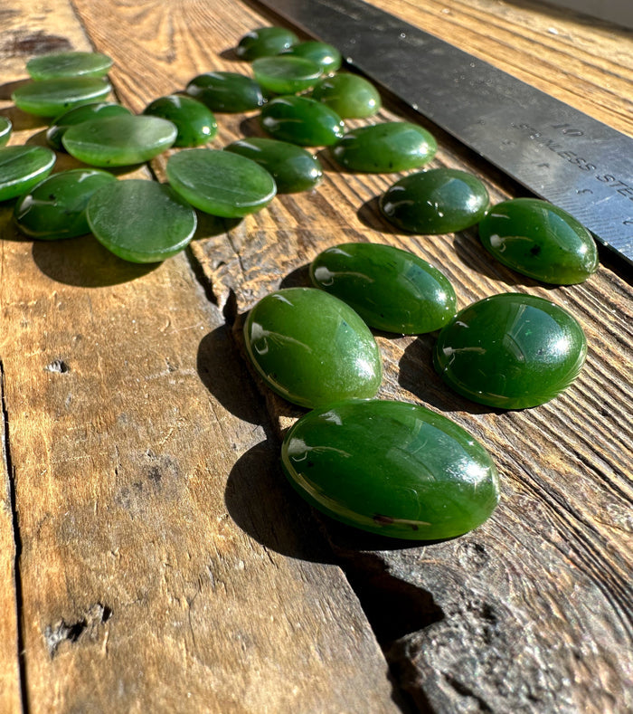 Close-up of glossy Canadian nephrite jade oval cabochons spread on wood with a metal ruler in the background for scale