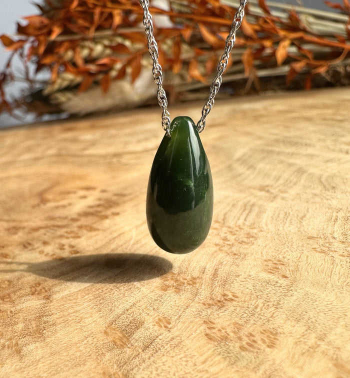 Dark green Canadian jade teardrop pendant suspended from a silver chain against a wooden surface.