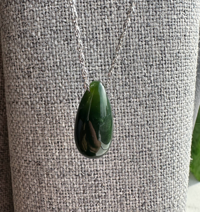 Dark green Canadian jade teardrop pendant on a silver chain resting against a gray woven fabric.