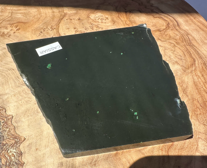 A dark green jade slab with irregular edges, featuring a small white label that reads 'CASSIAR', displayed on a richly grained wood surface.