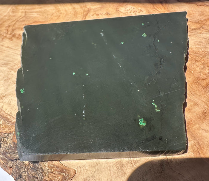 A square dark green jade slab with natural edge fractures and scattered small green inclusions, photographed on a wood surface.
