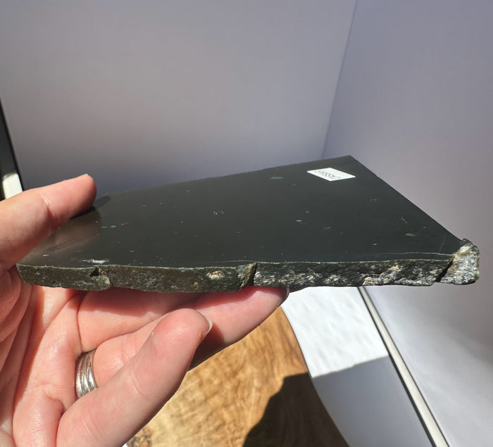 A hand holding a thick, polished JADE slab, showing its rough, textured natural edge. The slab has a small white sticker on one corner and is photographed in a casual indoor setting with a gray background and a wooden surface below.