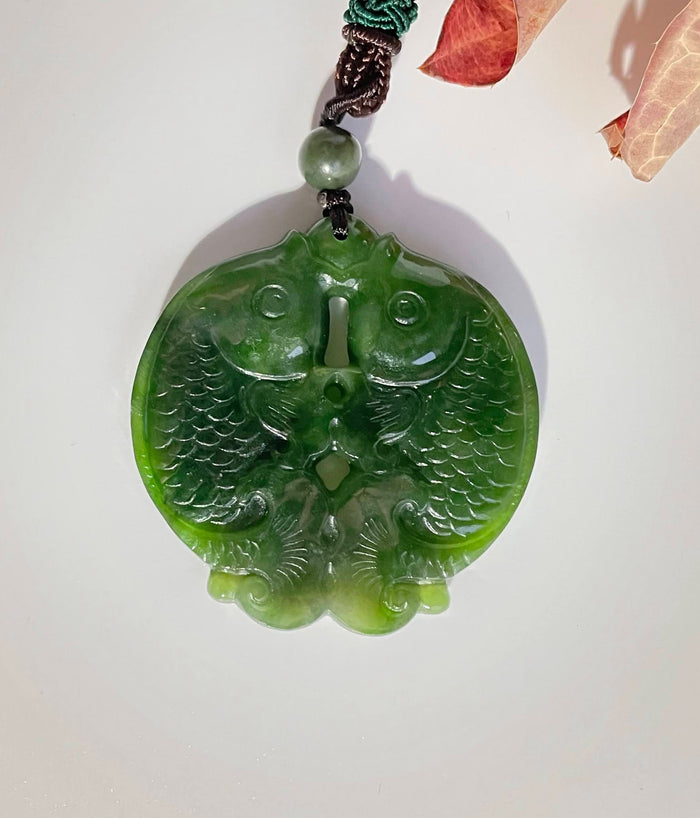 Green jade double koi fish pendant with carved detailing hanging from a cord.