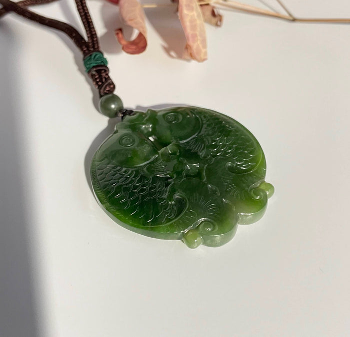 Circular green jade pendant carved with two koi fish facing each other and a small bead, on a brown cord.
