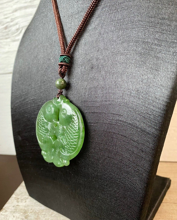 Green jade double koi fish pendant on a brown cord displayed on a black display bust.