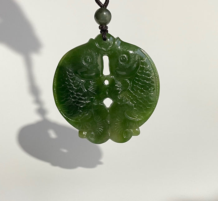 Close up of a glossy green circular jade pendant carved with two koi fish and swirling patterns, suspended from a brown cord.