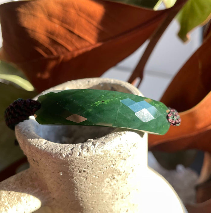 Faceted green jade bracelet with a dark maroon braided cord displayed on a textured ceramic vase.