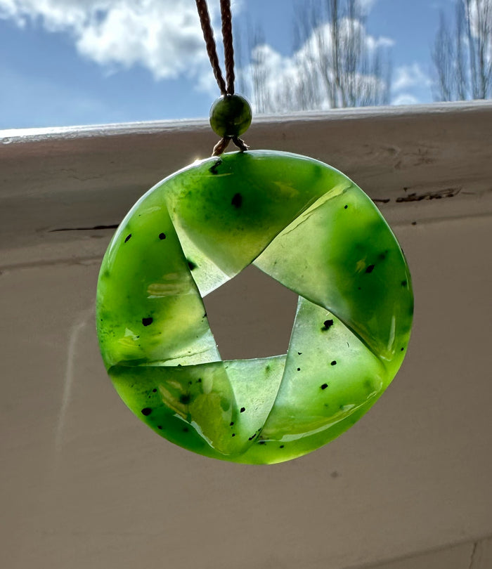 Green fractured AA nephrite jade pendant shaped as a circular wheel with four twisted segments speckled with black flecks.
