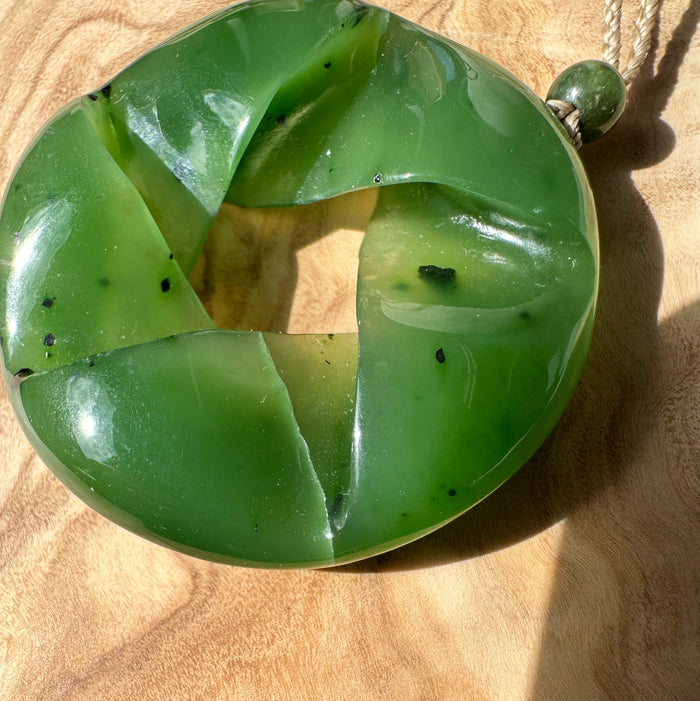 Close up of green nephrite jade circular pendant with four curved panels on a wooden surface, hanging by a rope and green bead.