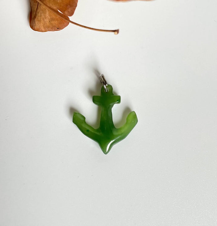 Bright green jade anchor charm with a metal loop on a white surface beside a dried leaf.
