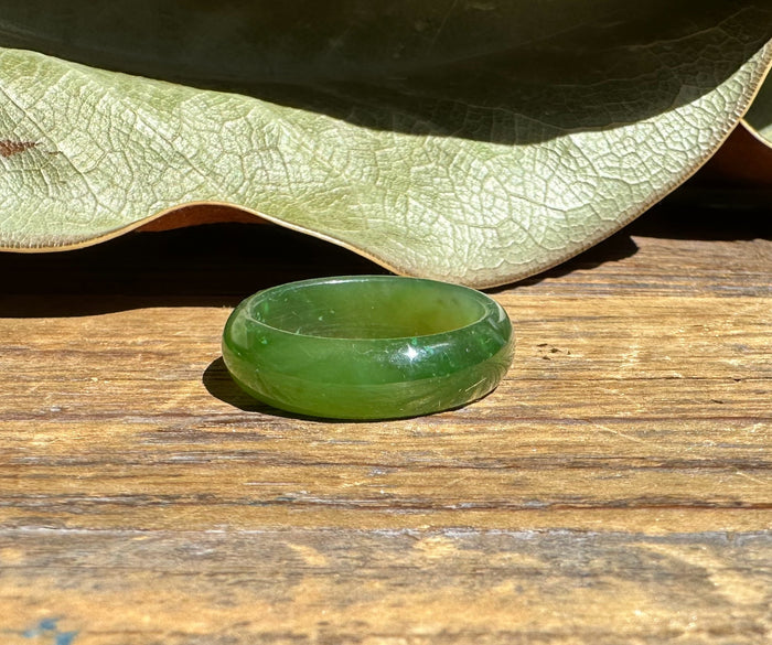 Single jade green band ring on a weathered wooden surface with a large leaf in the background.