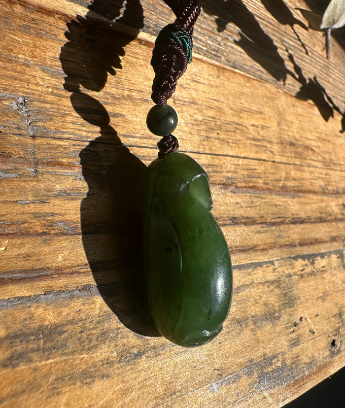 Green jade bird pendant on dark braided cord with a small green bead against a wooden background