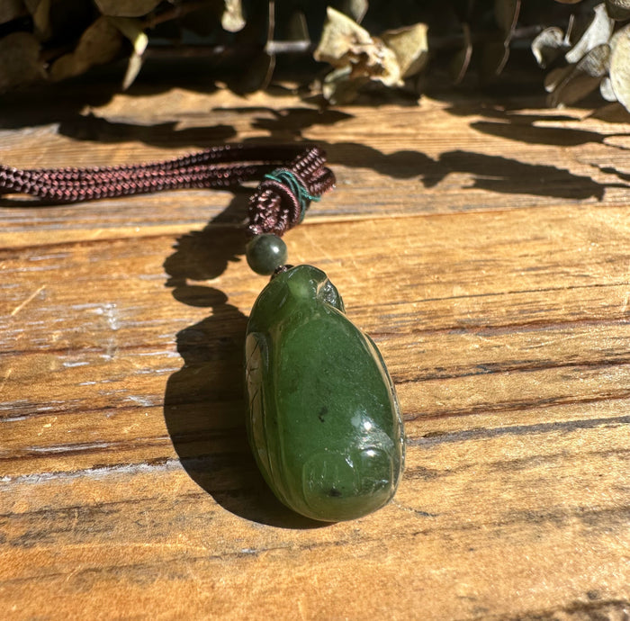 Dark green jade bird pendant with a round bead on a brown braided cord resting on a wooden surface