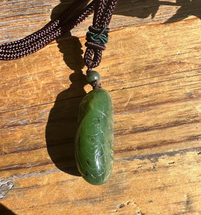 Green jade bird pendant on a brown braided cord resting on wood