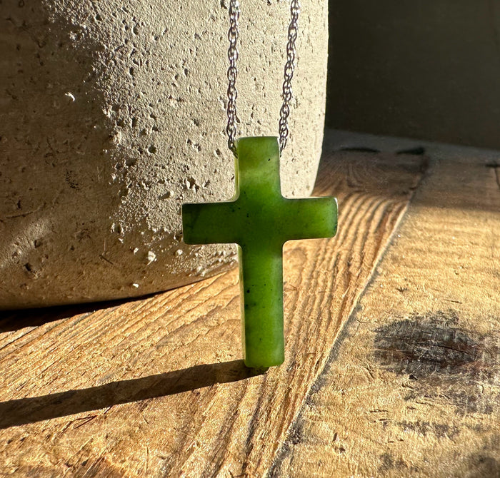 Bright green jade cross pendant on a silver chain against a textured stone and wood background.