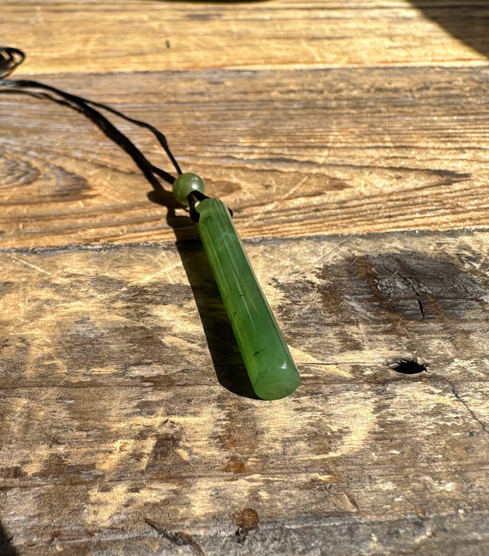 Green jade cylinder pendant with round bead above, suspended on a black cord against a wooden surface