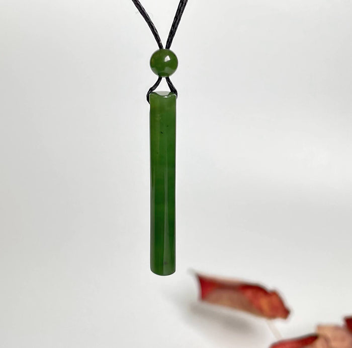 Green jade cylinder pendant on black cord with round bead, hanging against a white background