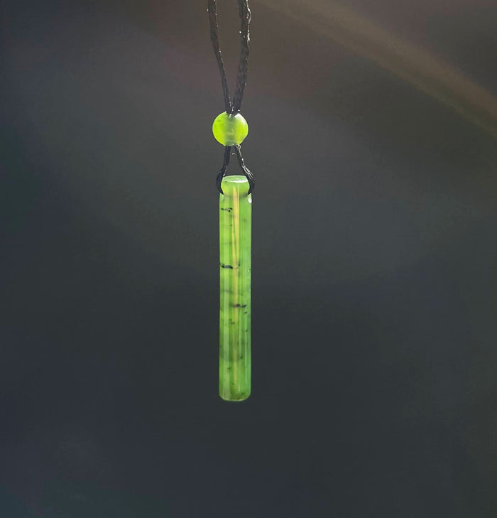 Jade cylinder pendant on a black cord with a round bead, shown against a dark blurred background
