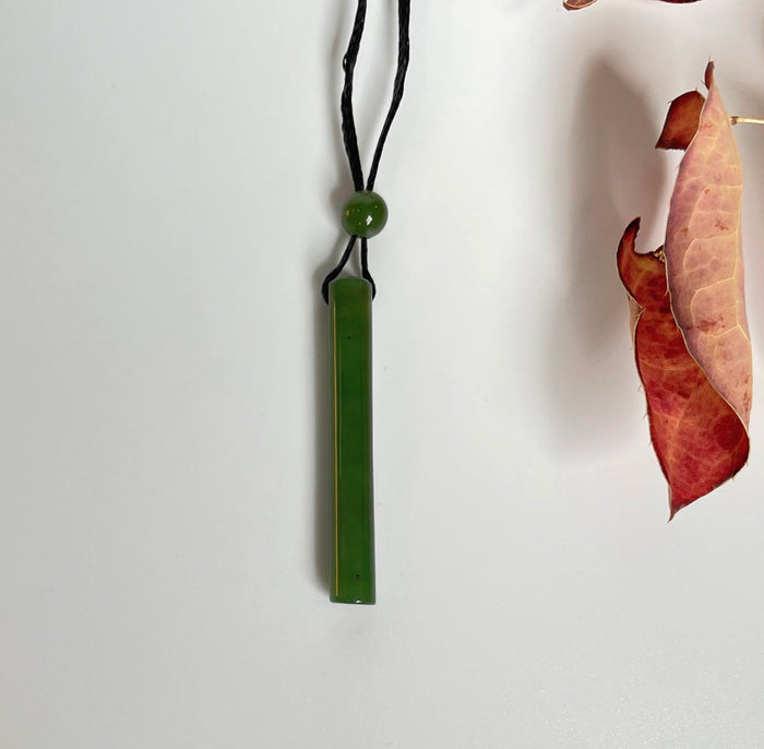 Green jade cylinder pendant on a black cord with a round bead, displayed against a white background