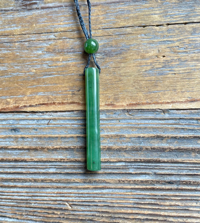 Green jade cylinder pendant on a black cord with a round bead resting on weathered wooden surface