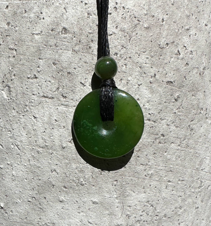 Jade donut pendant with a small bead on a black cord hanging against a textured stone wall.