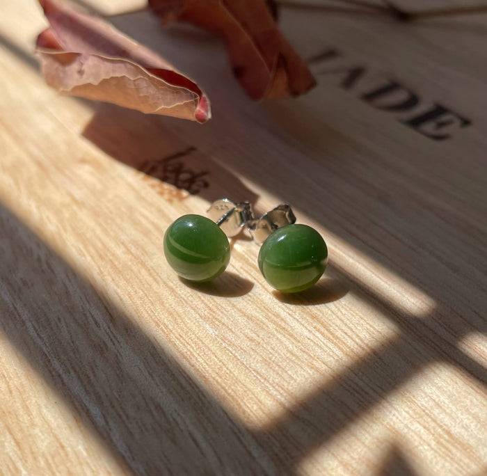 Green jade flat stud earrings with silver posts resting on a sunlit wooden surface.