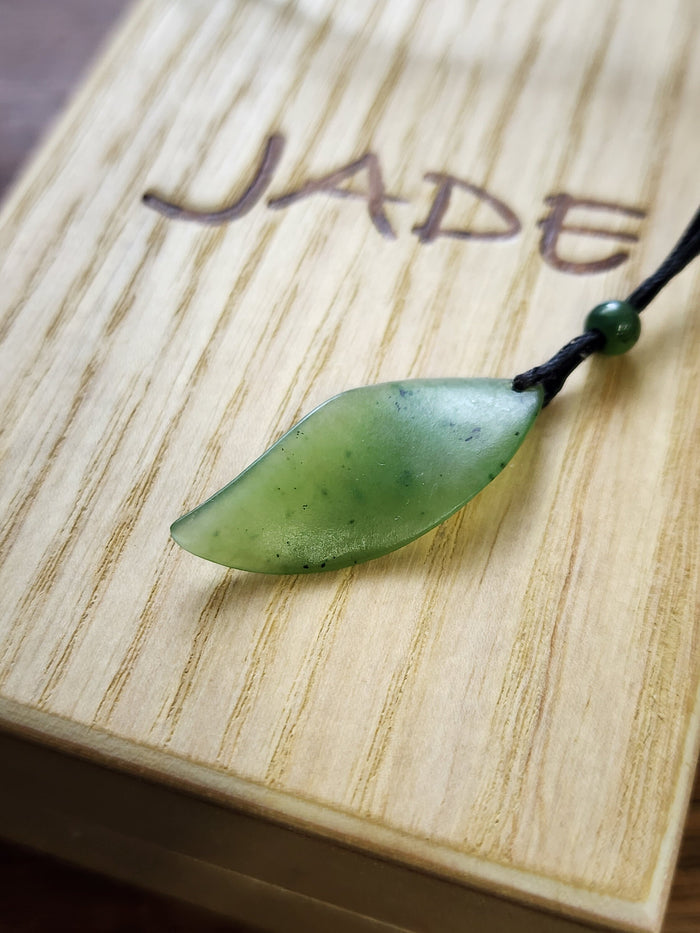 Pale green jade leaf pendant with dark specks on a black cord against a wood surface